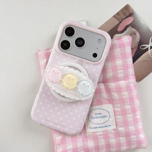 May include: A light pink smartphone case with a white polka dot pattern and a circular phone grip featuring three small, translucent, pastel-coloured ghost-like figures. The case has a cutout for a triple-lens camera system and Japanese text.