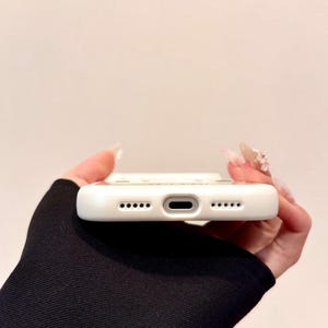 May include: A white phone case with a series of small holes and a central port. The case is held against a neutral background. The case has a smooth, clean surface and is designed to protect a smartphone.