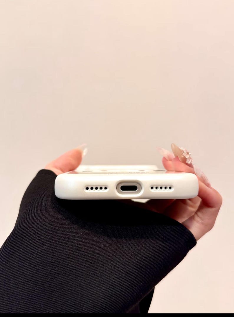 May include: A white phone case with a series of small holes and a central port. The case is held against a neutral background. The case has a smooth, clean surface and is designed to protect a smartphone.