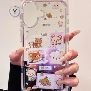 Puede incluir: Funda transparente para teléfono con diseño de oso Rilakkuma. La funda presenta varias ilustraciones del oso, incluyendo imágenes con texto en japonés. La funda se sostiene en una mano con uñas largas y decoradas.