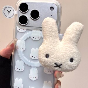 Op de afbeelding: Transparante telefoonhoes met een lichtblauwe achtergrond met witte konijnenillustraties en het woord "Miffy". Een pluizige witte konijnenkop is bevestigd aan de bovenkant van de hoes. De telefoonhoes heeft een zilveren cameramodule.