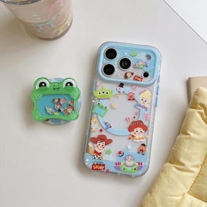 May include: A clear phone case featuring Toy Story character graphics and a light blue camera frame, paired with a green frog-shaped phone grip containing small floating character charms. The case displays Woody, Buzz Lightyear, and other icons.