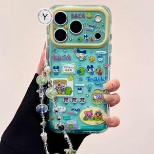 Puede incluir: Una funda transparente para tel&eacute;fono con un fondo turquesa y personajes de dibujos animados coloridos. La funda tiene un borde amarillo y una correa de cuentas con cuentas transparentes y verdes. Se ve texto japon&eacute;s.