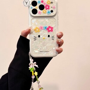 May include: White phone case featuring a Hello Kitty design with a floral crown and decorative flowers. The case has a pearlescent finish and includes a beaded wrist strap with a bow and additional floral accents.