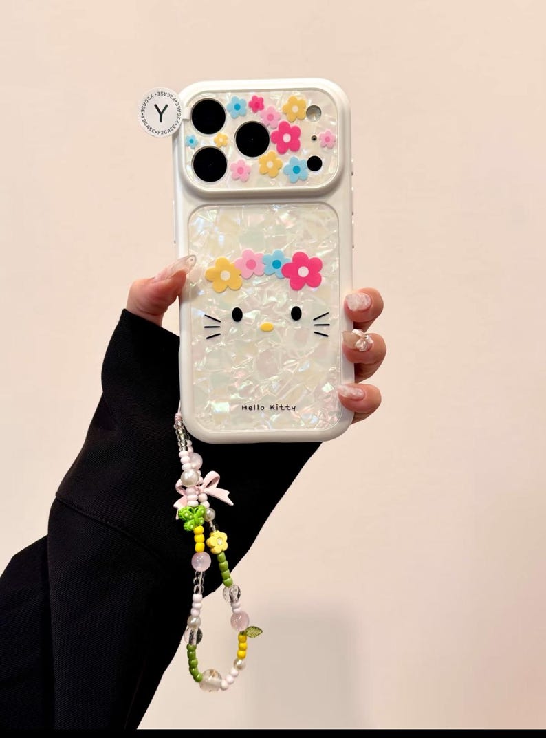 May include: White phone case featuring a Hello Kitty design with a floral crown and decorative flowers. The case has a pearlescent finish and includes a beaded wrist strap with a bow and additional floral accents.