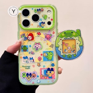 May include: A clear phone case with a green border, decorated with colorful Tamagotchi stickers. A matching Tamagotchi phone grip is attached. The case features various cartoon characters, stars, and musical notes. The text "TamaGoTchi" is visible.