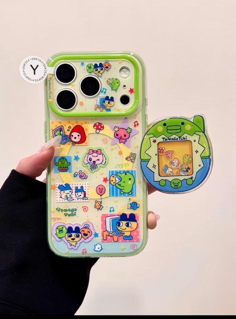 May include: A clear phone case with a green border, decorated with colorful Tamagotchi stickers. A matching Tamagotchi phone grip is attached. The case features various cartoon characters, stars, and musical notes. The text "TamaGoTchi" is visible.