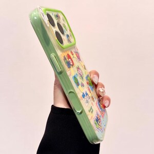May include: A green and clear phone case with a colorful cartoon design. The case has a green border and a clear back with various cartoon characters. The case is held up, showing the design and the phone's camera.
