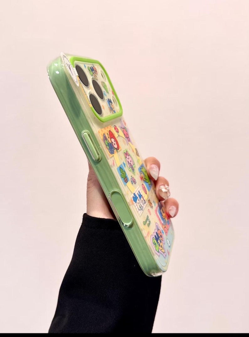 May include: A green and clear phone case with a colorful cartoon design. The case has a green border and a clear back with various cartoon characters. The case is held up, showing the design and the phone's camera.