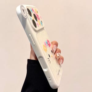 May include: White phone case with a floral design and a cartoon cat face. The case has a clear section with colorful flowers and a white border. The case has a sticker with the letter "Y".