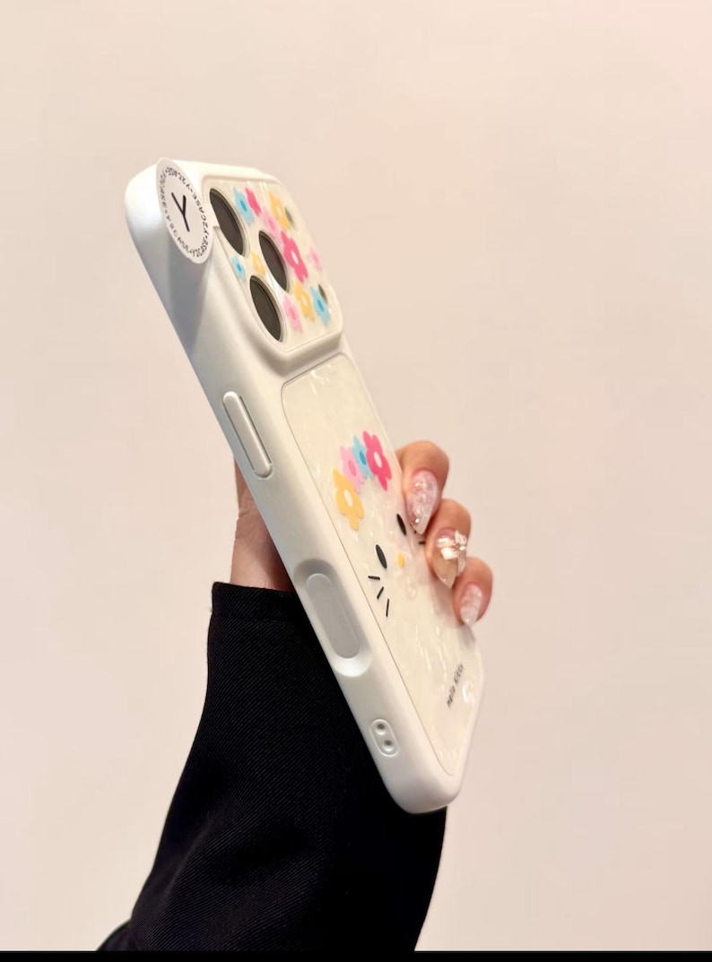 May include: White phone case with a floral design and a cartoon cat face. The case has a clear section with colorful flowers and a white border. The case has a sticker with the letter "Y".