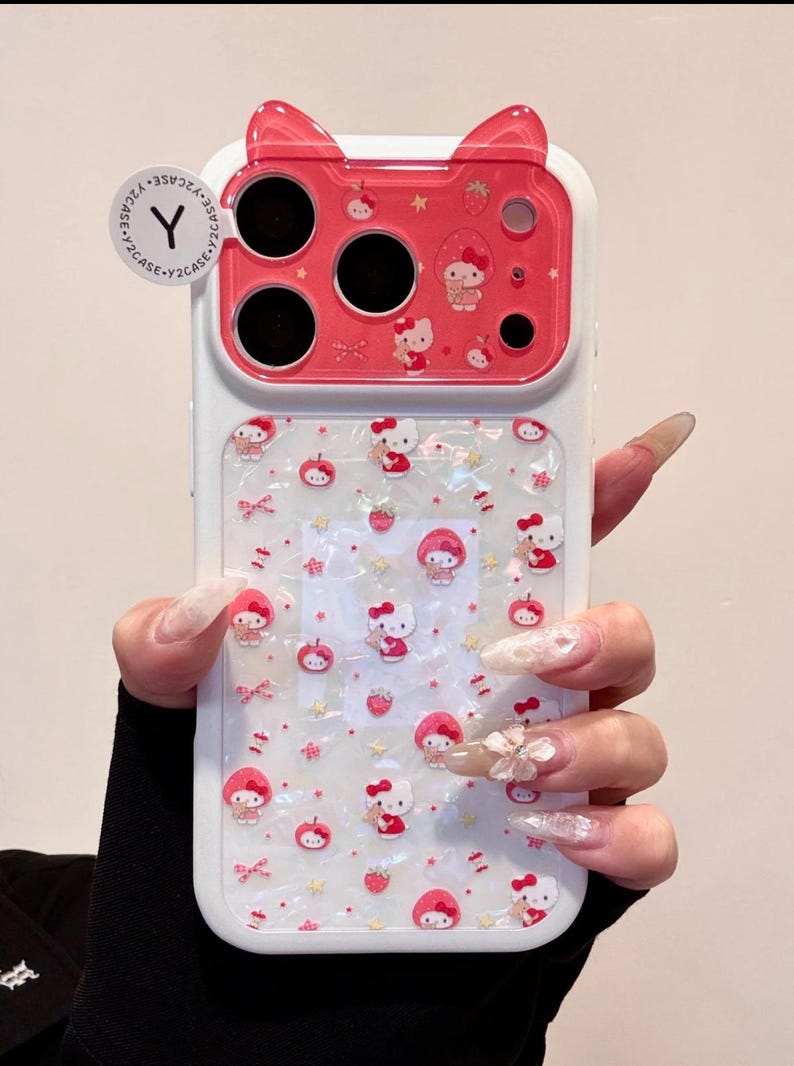 A hand holds a Hello Kitty phone case with a red cat-ear camera section and a pearly white patterned back.