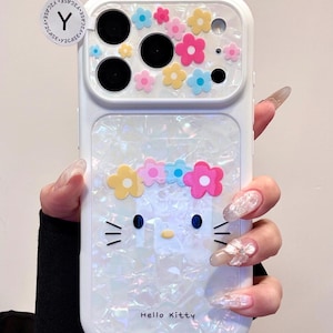 May include: White phone case featuring a Hello Kitty design with a flower crown and colorful flower accents. The case has a pearlescent finish and the text "Hello Kitty" is visible.