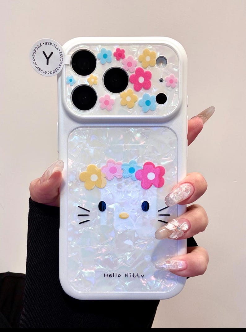 May include: White phone case featuring a Hello Kitty design with a flower crown and colorful flower accents. The case has a pearlescent finish and the text "Hello Kitty" is visible.