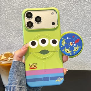 May include: A lime green smartphone case featuring the three-eyed alien character from Toy Story, with a purple and blue belt design at the bottom. A circular blue phone grip with alien faces, stars, and a rocket ship is attached to the side.