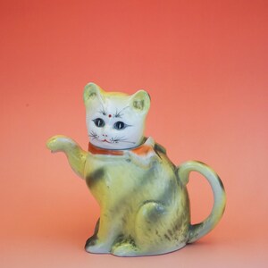 Op de afbeelding: Een gele en grijze keramische katten theepot met een witte kop en een oranje kraag. De theepot heeft een handvat en een tuit. Het gezicht van de kat heeft blauwe ogen en een rode stip. Perzik achtergrond.