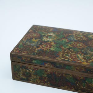 May include: A rectangular metal box featuring a detailed cloisonné enamel surface with a dense pattern of multi-colored flowers and leaves in shades of red, yellow, green, and blue, set against a dark background with visible metal outlines.