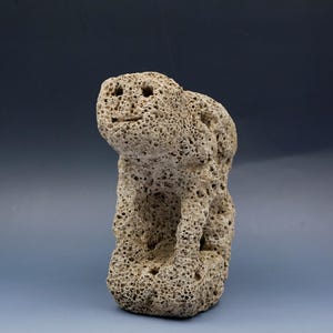 Può includere: Una scultura in pietra beige e porosa a forma di creatura seduta. La figura presenta una testa arrotondata con due piccole rientranze circolari per gli occhi e una linea orizzontale per la bocca. La superficie è molto porosa e irregolare.