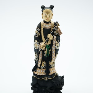 May include: A detailed figurine of a person in traditional Asian attire. The figure is predominantly black with gold and white accents, holding a staff. The face and hair are gold. The figure stands atop a black creature.