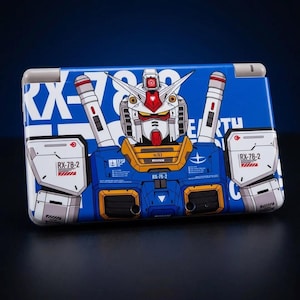 May include: A blue and white case featuring a detailed illustration of a Gundam robot. The design includes the robot's face and body, with the text "RX-78-2" and other Japanese text. The case has a rectangular shape with rounded corners.