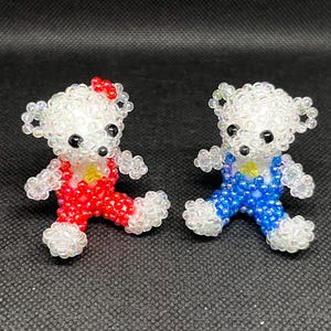 May include: Two beaded teddy bear figurines. One bear is wearing red overalls and a red bow, the other is wearing blue overalls. Both bears have white bodies, black eyes, and yellow accents.
