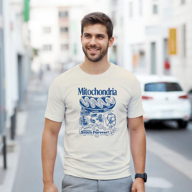 A man with short brown hair and a beard smiles while wearing a cream-colored t-shirt with a blue mitochondria graphic, standing on a city street.