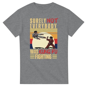 Puede incluir: Una camiseta gris jaspeado de manga corta con un estampado gráfico de dos figuras pixeladas en poses de artes marciales. El texto dice SURELY NOT EVERYBODY WAS KUNG FU FIGHTING, sobre un fondo de atardecer con rayas horizontales en amarillo, naranja, rojo y azul.