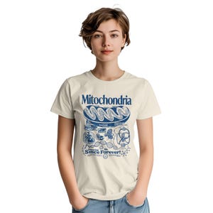 A woman with short brown hair stands facing forward, wearing a cream-colored t-shirt with a blue mitochondria graphic.