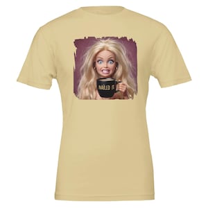 Op de afbeelding: Een lichtbeige t-shirt met korte mouwen en een grafische print van een blonde pop met een wijdopen blik en ontblote tanden die een zwarte mok vasthoudt. De mok heeft de tekst NAILED IT in gouden letters tegen een donkerpaarse vierkante achtergrond.