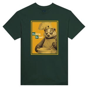 Puede incluir: Camiseta verde oscuro con un gráfico humorístico. Representa un tejón con gorro de chef y delantal, sosteniendo un cuenco y una cuchara. El gráfico incluye el texto "Baking Bread" y un símbolo de la tabla periódica.