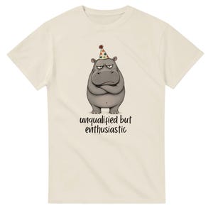 Peut inclure: Un t-shirt à manches courtes de couleur crème avec un motif représentant un hippopotame gris portant un chapeau de fête à pois colorés, les bras croisés. Sous l'hippopotame, le texte "unqualified but enthusiastic" est écrit en noir.