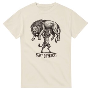 Könnte beinhalten: Ein cremefarbenes Kurzarm-T-Shirt mit einer schwarz-weißen grafischen Illustration eines Zebras, das einen Löwen über seinen Kopf hebt. Unter der Illustration ist der Text BUILT DIFFERENT in einer fetten, dunklen Schriftart gedruckt.