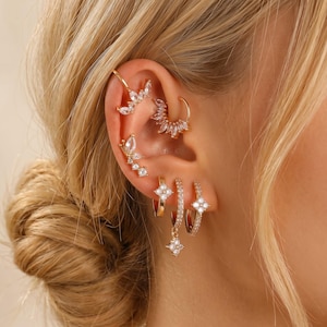 May include: A selection of gold-coloured earrings and ear cuffs, embellished with clear crystals. The ear cuffs showcase leaf and petal designs, while the earrings include hoop and drop styles with floral accents. The jewellery is set against a backdrop of blonde hair.