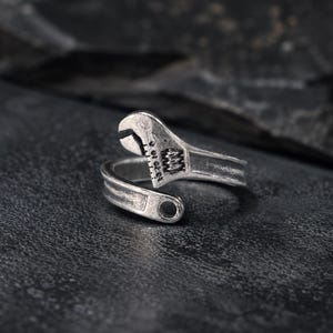 Puede incluir: Un anillo ajustable de tono plateado con forma de llave inglesa. La banda presenta un acabado metálico texturizado y desgastado, con una representación realista de la cabeza de una llave y una pequeña abertura circular en el extremo opuesto.