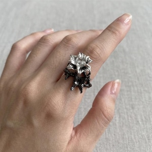 May include: A silver and black floral ring. The ring features a detailed, three-dimensional flower design with a contrasting color scheme. The ring is worn on a finger, showcasing its intricate details and unique style. The ring is a piece of jewelry.