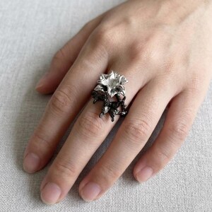 May include: A silver-colored ring featuring a floral design. The ring has a two-tone appearance, with the flower petals in silver and the band and stems in black. The ring is worn on a finger.