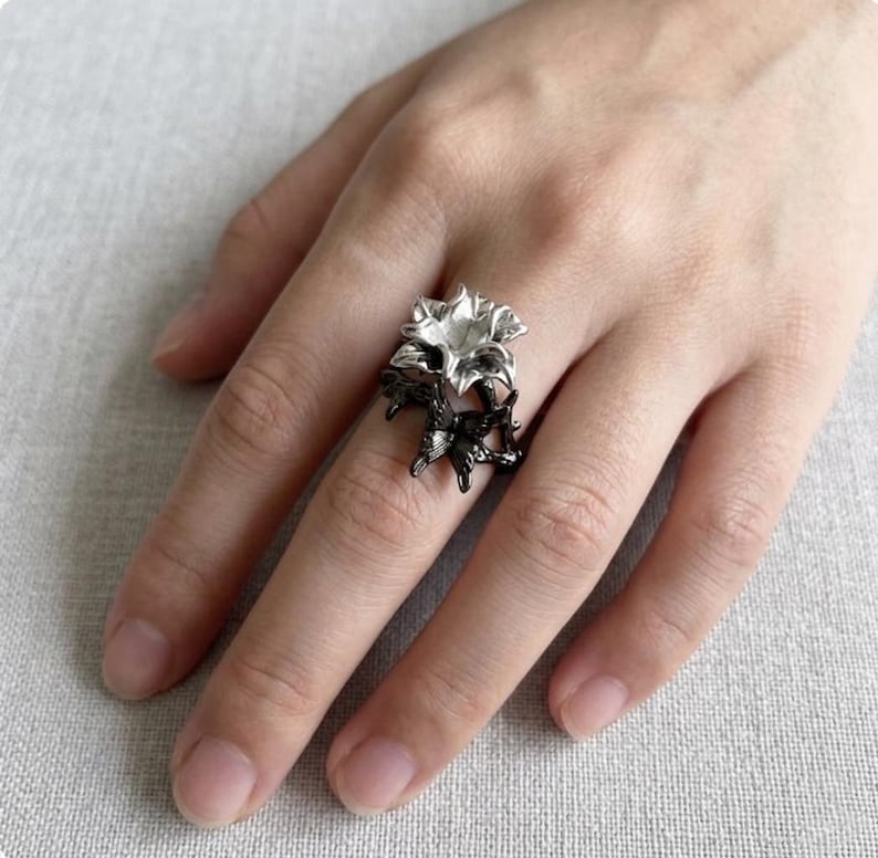 May include: A silver-colored ring featuring a floral design. The ring has a two-tone appearance, with the flower petals in silver and the band and stems in black. The ring is worn on a finger.