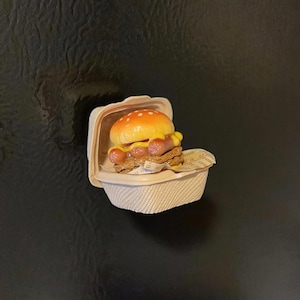 Puede incluir: Una hamburguesa en miniatura con dos perritos calientes, mostaza y un panecillo de sésamo, presentada en un pequeño recipiente beige para llevar. El alimento descansa sobre un trozo de papel impreso. La estética general es una miniatura lúdica con temática de comida.