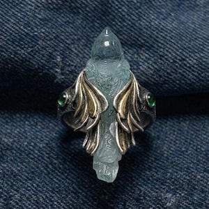 Puede incluir: Un anillo de plata detallado con un pájaro de piedra preciosa azul claro tallado con las alas extendidas. Las alas tienen detalles dorados y el anillo está adornado con dos gemas verdes a cada lado del pájaro.