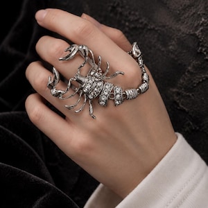 May include: A silver-toned scorpion ring featuring articulated segments that extend across the finger. The body and tail of the scorpion are encrusted with clear, sparkling rhinestones, while the pincers and legs are crafted from polished metal.