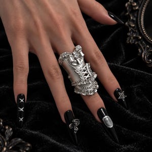 Könnte beinhalten: Ein silberfarbener Ring, der den gesamten Finger bedeckt und wie ein Korsett geformt ist, mit gerüschten Kanten, einer zentralen Schleife und Schnürdetails. Der Ring sitzt am Mittelfinger und zeigt ein metallisches Finish mit Texturen.