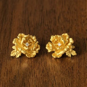 May include: A pair of gold-colored flower-shaped stud earrings. Each earring features detailed petals and a small, sparkling gemstone in the center. The earrings are displayed on a wooden surface, showcasing their intricate design and craftsmanship.