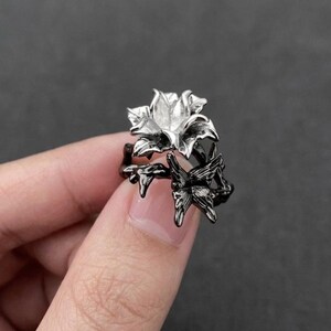 May include: A silver-colored flower ring with a dark gray band and butterfly accents. The ring features a detailed floral design with a butterfly on the side. The ring is a piece of jewelry.