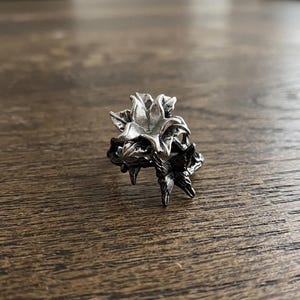 May include: A silver-colored ring with a floral design. The ring features a detailed flower with multiple petals and leaves. The ring is set against a wooden surface.