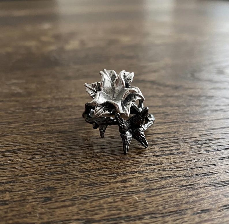 May include: A silver-colored ring with a floral design. The ring features a detailed flower with multiple petals and leaves. The ring is set against a wooden surface.