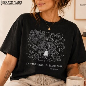 Może przedstawiać: Czarny t-shirt z białą grafiką przedstawiającą osobę przytłoczoną otwartymi kartami komputera z etykietami „Praca”, „ADHD”, „Memy” i innymi. Tekst brzmi „47 KART OTWARTYCH. 0 ZADAŃ WYKONANYCH. JEST OK.” Koszulka zawiera również tekst „BRAIN TABS - Zamykanie kart mentalnych od 2020 r.”