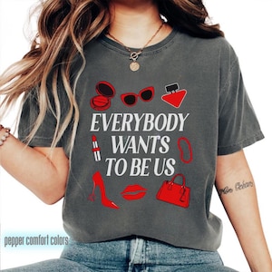 Puede incluir: Camiseta gris oscuro con el texto "EVERYBODY WANTS TO BE US" e ilustraciones rojas de maquillaje, gafas de sol, perfume, lápiz labial, una pulsera, un tacón alto, labios y un bolso. La camiseta tiene cuello redondo.