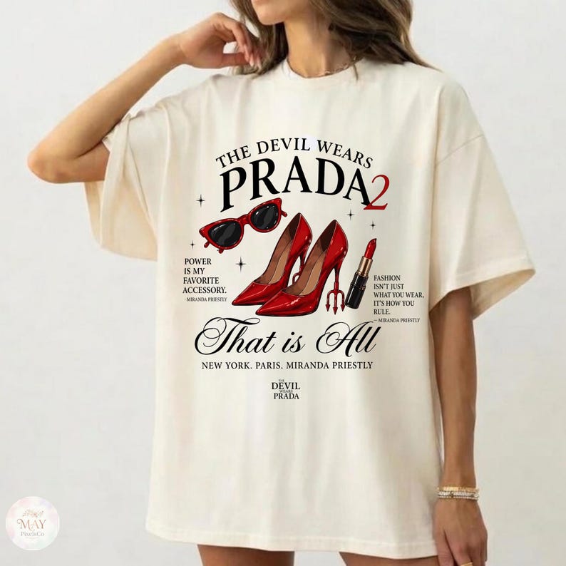 May include: Cream-colored t-shirt with a graphic design featuring the text "THE DEVIL WEARS PRADA 2". The design includes red high heels, sunglasses, and lipstick. Additional text includes "That is All" and "Power is my favorite accessory."