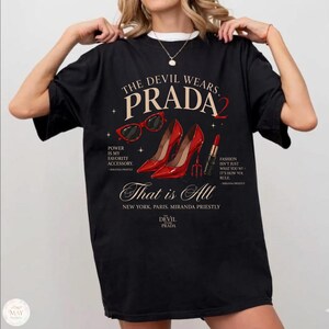 May include: Black t-shirt with the text "THE DEVIL WEARS PRADA 2" and fashion-related graphics. The design includes red high heels, sunglasses, and lipstick, with quotes about fashion and accessories. The shirt also has the text "That is All".