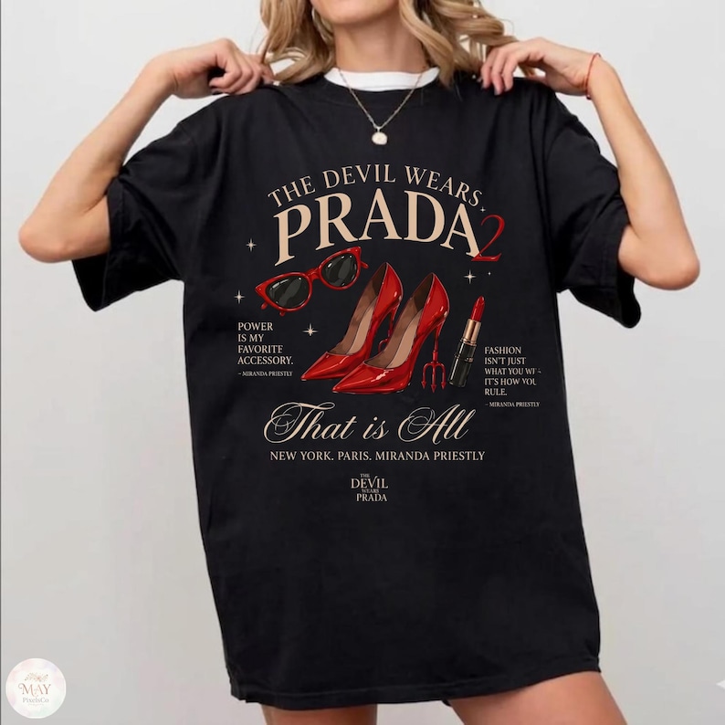 May include: Black t-shirt with the text "THE DEVIL WEARS PRADA 2" and fashion-related graphics. The design includes red high heels, sunglasses, and lipstick, with quotes about fashion and accessories. The shirt also has the text "That is All".